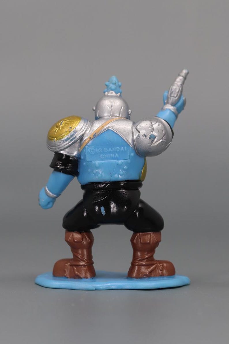May include: A blue, silver, and black action figure with a gun raised in the air. The figure has brown boots, black pants, and a blue torso. The figure is standing on a light blue base. The figure has gold and silver shoulder pads.