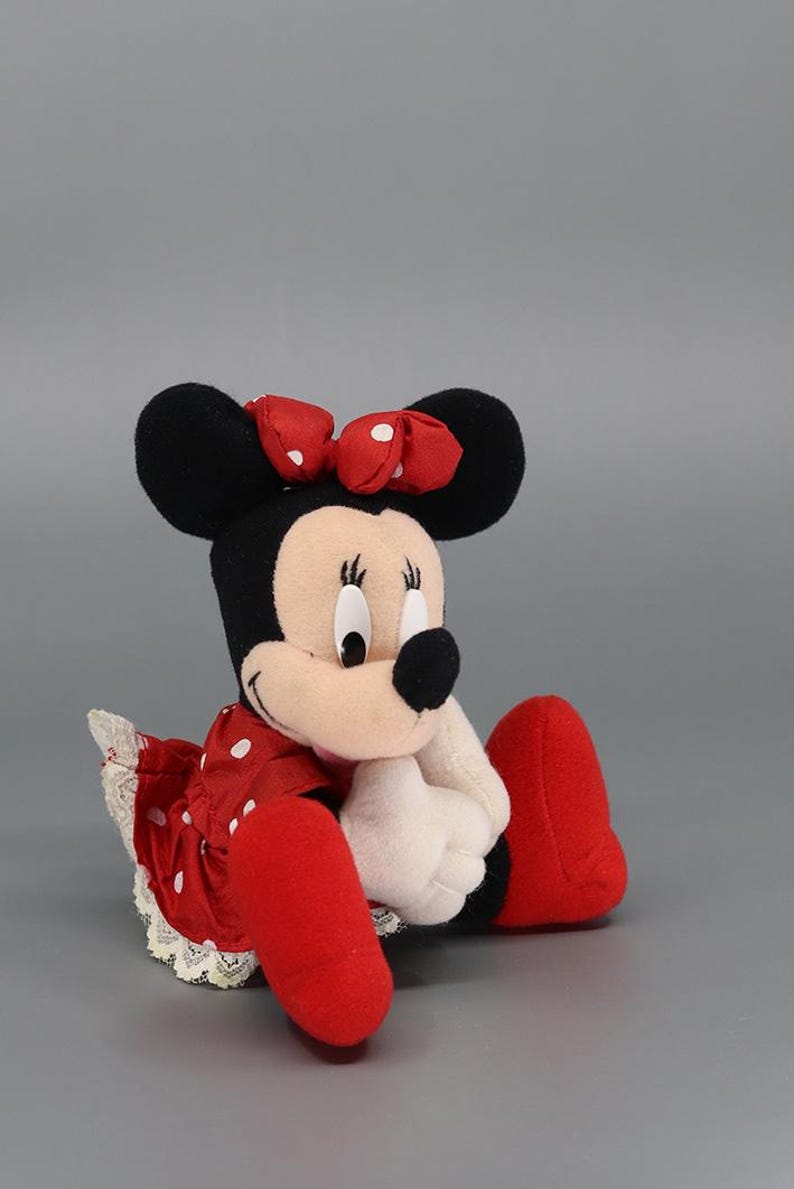 May include: A plush Minnie Mouse doll, featuring black ears, a red polka-dot bow, and a red dress with white polka dots. The doll has white gloves and red shoes. The doll is sitting down.