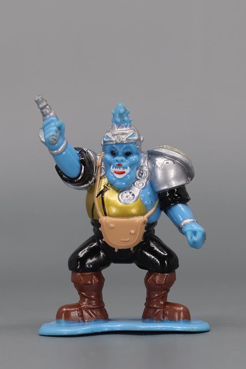 May include: A blue action figure with a silver helmet and shoulder pads, black pants, and brown boots. The figure has a gold chest and a tan satchel. It is holding a silver weapon and standing on a blue base.