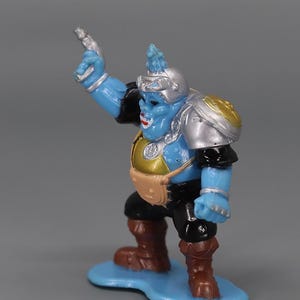 May include: A blue action figure with silver and gold armor, black sleeves, and brown boots. The figure is standing on a blue base and holding a silver weapon. The figure has a helmet and a small pouch.