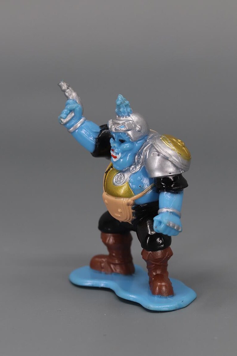 May include: A blue action figure with silver and gold armor, black sleeves, and brown boots. The figure is standing on a blue base and holding a silver weapon. The figure has a helmet and a small pouch.