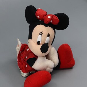 May include: A plush Minnie Mouse doll, featuring black ears, a red bow with white polka dots, and a red dress with white polka dots. The doll has white gloves, a black nose, and red shoes. The doll is sitting with its legs extended.