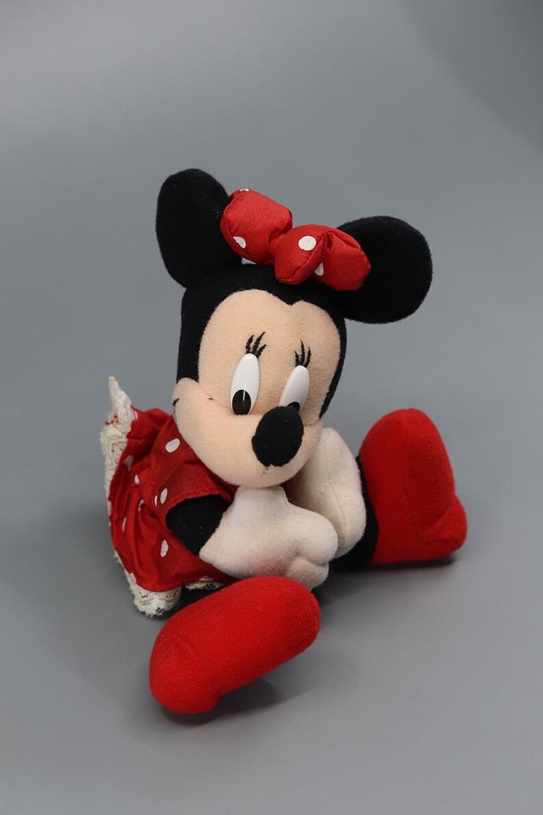May include: A plush Minnie Mouse doll, featuring black ears, a red bow with white polka dots, and a red dress with white polka dots. The doll has white gloves, a black nose, and red shoes. The doll is sitting with its legs extended.