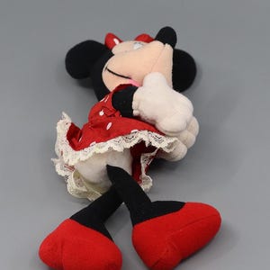 May include: A Minnie Mouse plush toy, featuring black ears, a red bow, and a red dress with white polka dots. The dress has a white lace trim. The toy has white gloves and red shoes. The plush is lying on its side.