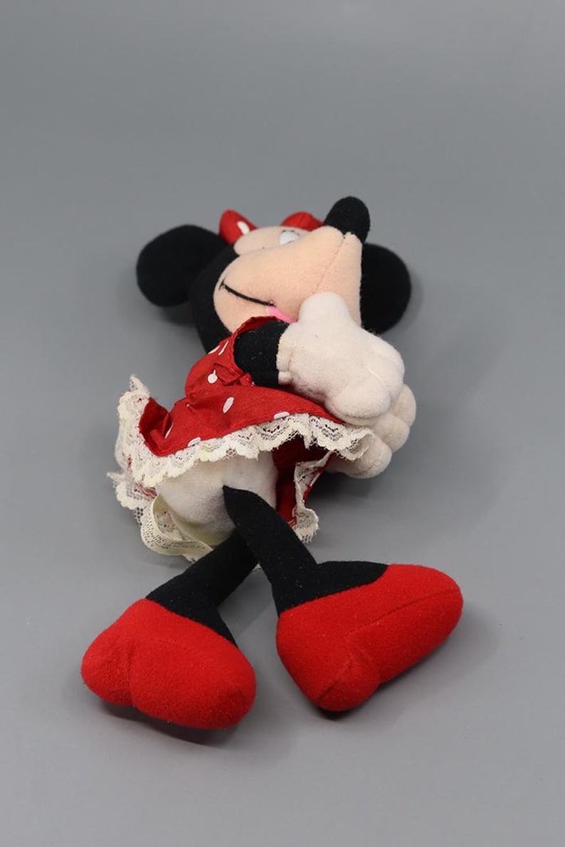 May include: A Minnie Mouse plush toy, featuring black ears, a red bow, and a red dress with white polka dots. The dress has a white lace trim. The toy has white gloves and red shoes. The plush is lying on its side.