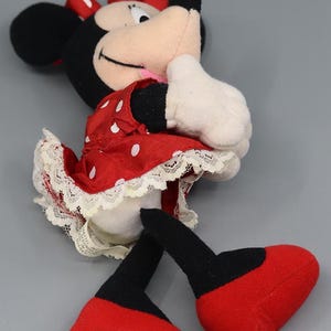 May include: A plush Minnie Mouse doll, featuring a red dress with white polka dots and a matching bow. The doll has black ears, a pink tongue, and white gloves. The doll's feet are red, and the legs are black.