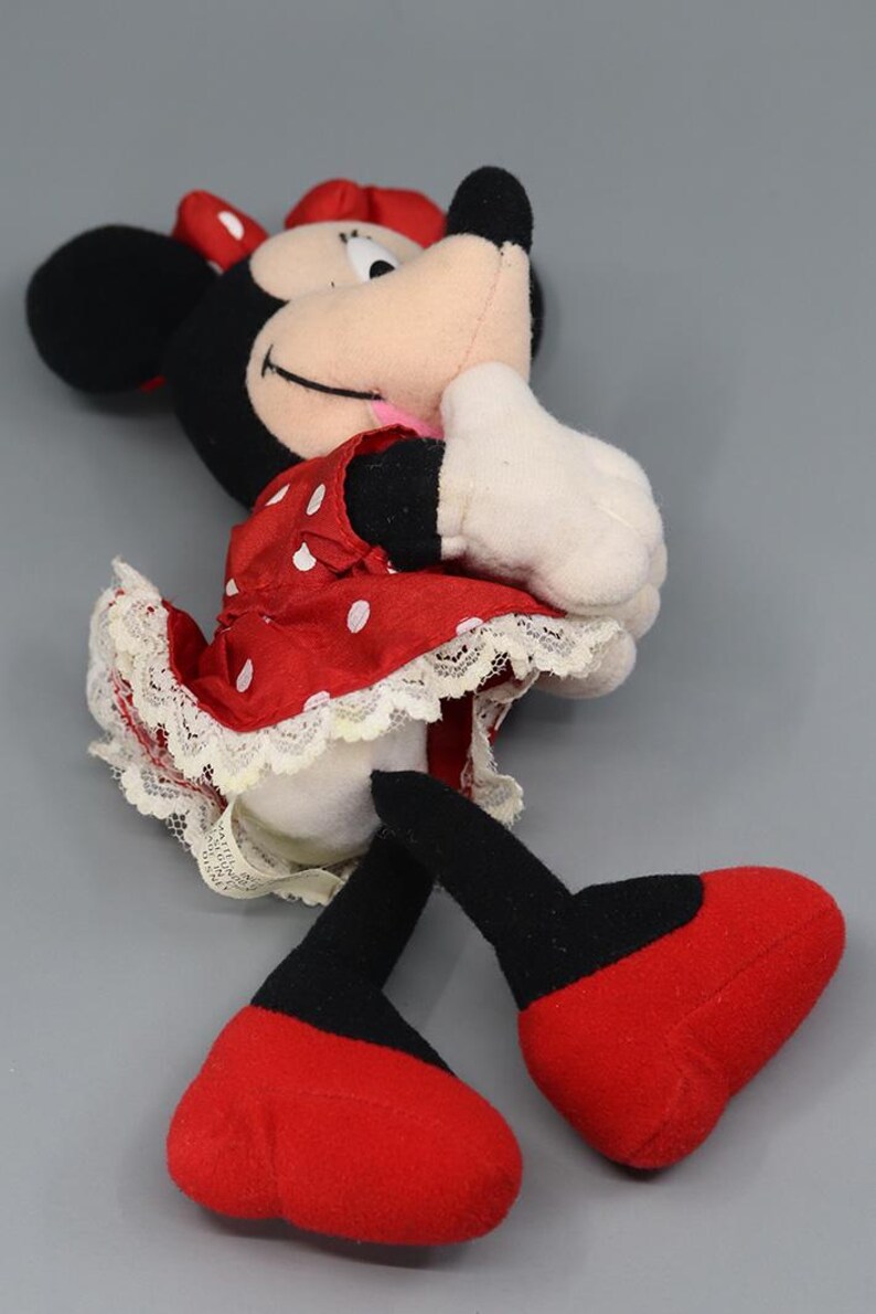May include: A plush Minnie Mouse doll, featuring a red dress with white polka dots and a matching bow. The doll has black ears, a pink tongue, and white gloves. The doll's feet are red, and the legs are black.