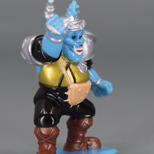 May include: A blue action figure with a silver helmet and shoulder pads, a gold torso, black pants, and brown boots. The figure has a tan satchel and is standing on a blue base. The figure is holding its right arm up.