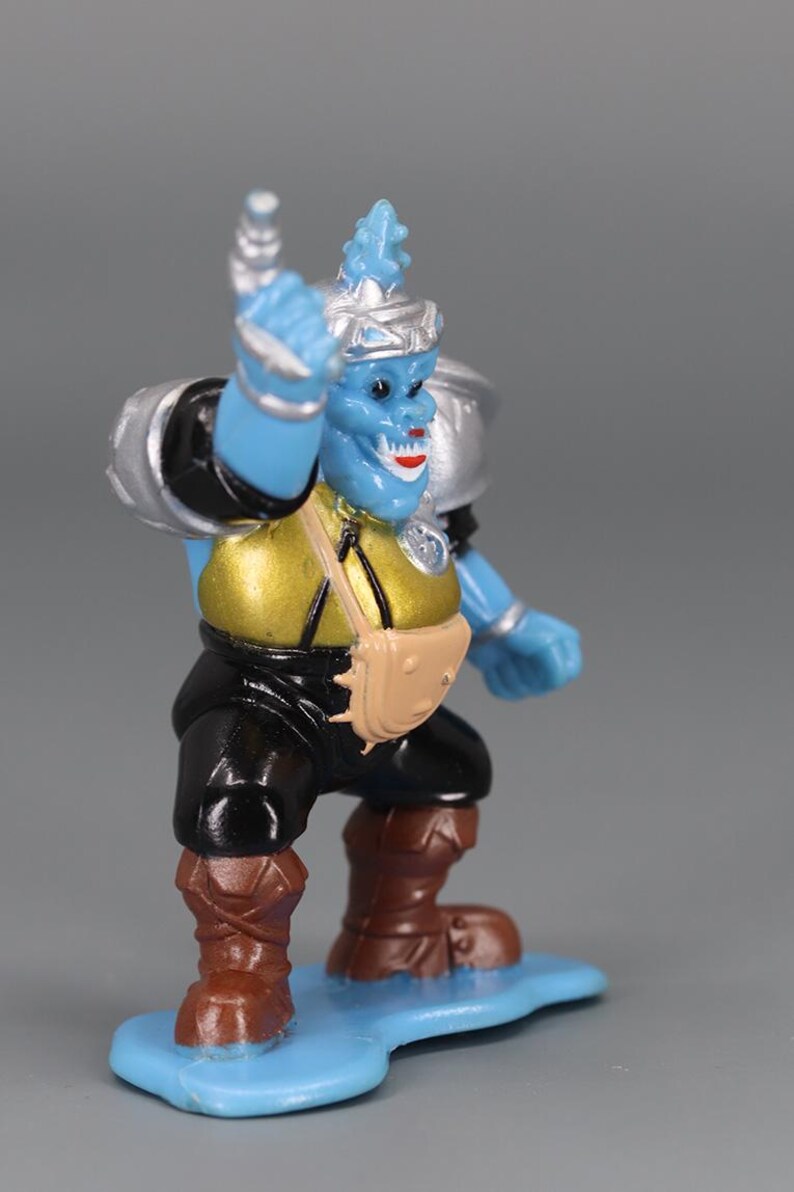 May include: A blue action figure with a silver helmet and shoulder pads, a gold torso, black pants, and brown boots. The figure has a tan satchel and is standing on a blue base. The figure is holding its right arm up.