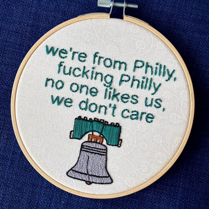 "we're from Philly, fucking Philly, no one likes us, we don't care". Quote hand embroidered in eagles green. Silver, green, black liberty bell below. Sewn on white-on-white cotton. Displayed in light wooden embroidery hoop as wall art.
