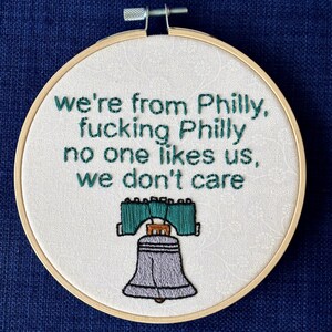 "we're from Philly, fucking Philly, no one likes us, we don't care". Quote hand embroidered in eagles green. Silver, green, black liberty bell below. Sewn on white-on-white cotton. Displayed in light wooden embroidery hoop as wall art.