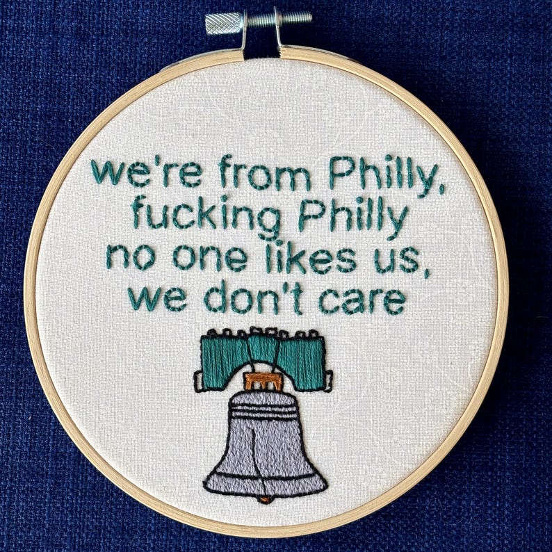 "we're from Philly, fucking Philly, no one likes us, we don't care". Quote hand embroidered in eagles green. Silver, green, black liberty bell below. Sewn on white-on-white cotton. Displayed in light wooden embroidery hoop as wall art.