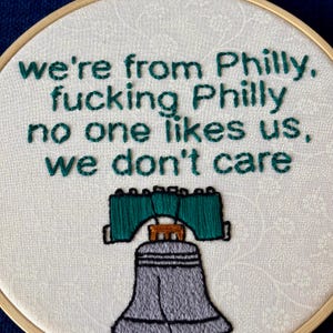 "we're from Philly, fucking Philly, no one likes us, we don't care". Quote hand embroidered in eagles green. Silver, green, black liberty bell below. Sewn on white-on-white cotton. Displayed in light wooden embroidery hoop as wall art.