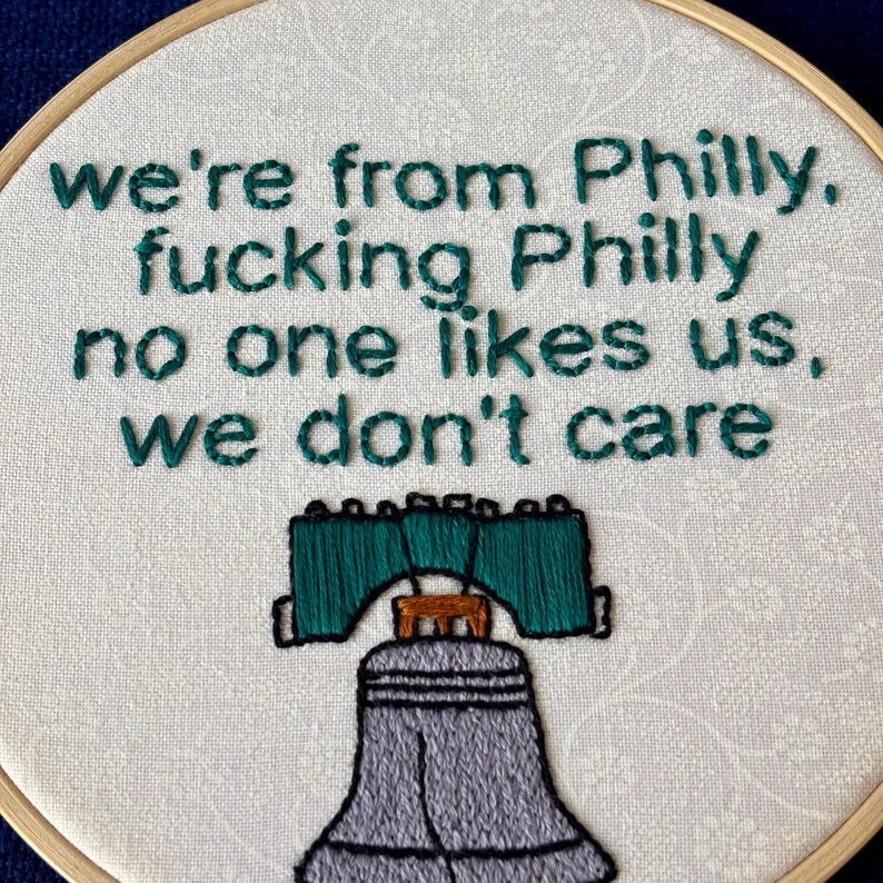 "we're from Philly, fucking Philly, no one likes us, we don't care". Quote hand embroidered in eagles green. Silver, green, black liberty bell below. Sewn on white-on-white cotton. Displayed in light wooden embroidery hoop as wall art.