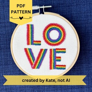 May include: A circular wooden embroidery hoop containing white fabric with the word LOVE stitched in rainbow-colored thread. The letter O is a circular rainbow shape, while the L, V, and E are formed with vertical rainbow stripes.