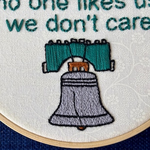 "we're from Philly, fucking Philly, no one likes us, we don't care". Quote hand embroidered in eagles green. Silver, green, black liberty bell below. Sewn on white-on-white cotton. Displayed in light wooden embroidery hoop as wall art.