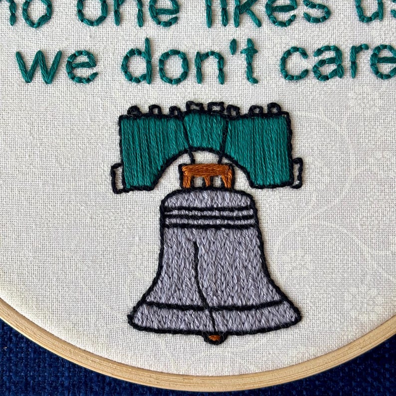 "we're from Philly, fucking Philly, no one likes us, we don't care". Quote hand embroidered in eagles green. Silver, green, black liberty bell below. Sewn on white-on-white cotton. Displayed in light wooden embroidery hoop as wall art.
