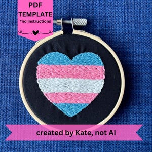 May include: An embroidery hoop with a black fabric background features a heart design in the colors of the transgender flag: light blue, pink, and white. The text "PDF TEMPLATE *no instructions" is visible. The text "created by Kate, not AI" is also visible.