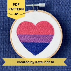 May include: An embroidered heart in the colors of the bisexual pride flag, displayed in a wooden embroidery hoop. The heart is divided into three sections: pink, purple, and blue. Text on the image reads "PDF PATTERN" and "created by Kate, not AI."