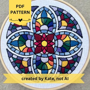 May include: A circular embroidery hoop containing a white fabric panel with a colourful, symmetrical stained-glass style pattern. The design features a central red and yellow flower surrounded by segments of blue, green, purple, and orange thread.