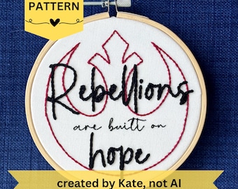 Op de afbeelding: Een ronde houten borduurring met witte stof waarop een rood geborduurd sterrenvogel-symbool staat. De zwarte geborduurde tekst luidt "Rebellions are built on hope". Het ontwerp is gecentreerd binnen het frame van de borduurring.