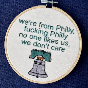 "we're from Philly, fucking Philly, no one likes us, we don't care". Quote hand embroidered in eagles green. Silver, green, black liberty bell below. Sewn on white-on-white cotton. Displayed in light wooden embroidery hoop as wall art.