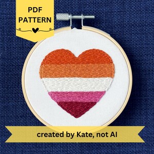 May include: An embroidery hoop with a heart design in the colors of the lesbian pride flag. The heart is stitched in orange, white, pink, and burgundy thread. The text "PDF PATTERN" is in a yellow banner, and the text "created by Kate, not AI" is in a yellow banner.