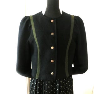 Könnte beinhalten: Schwarze Wolljacke mit rundem Ausschnitt und langen Ärmeln. Die Jacke hat olivgrüne Paspeln und goldfarbene Knöpfe. Die Jacke wird auf einer schwarzen Schaufensterpuppe präsentiert.