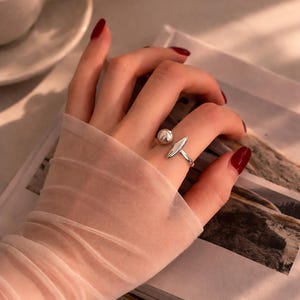 May include: A silver ring with a spherical and elongated oval design. The ring is worn on a finger with red nail polish. The hand is covered with a sheer, light pink fabric. A cup of coffee and a book are in the background.
