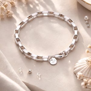 May include: A silver bracelet with a rectangular link design. The bracelet is made of polished metal and features a lobster clasp closure. A small, round charm with the number "925" is attached. The jewelry is displayed on a light-colored surface.