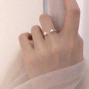 May include: A silver ring with an open design, featuring a small square and a sphere on either side of the opening. The ring is worn on a finger, with a book in the background. The ring is a simple, modern piece of jewelry.
