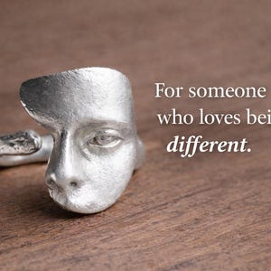 May include: A silver-colored ring with a face design. The ring features a detailed depiction of a human face. The text on the image reads: "For someone who loves being different."