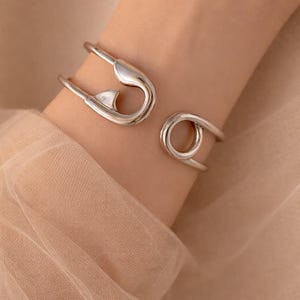 May include: A silver bracelet with a safety pin design. The bracelet has a large safety pin shape and a circular design. The jewellery is worn on a wrist, with a soft, neutral-toned background.