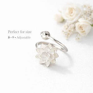 May include: A silver-toned ring featuring a white flower design. The ring is adjustable and is designed for sizes 8-9. The ring is set against a white background with white flowers in the background.