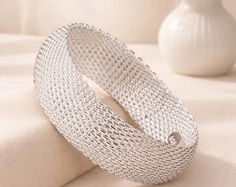 May include: A silver-colored bracelet with a woven mesh design. The bracelet is a wide band, and the mesh pattern creates a textured surface. The bracelet is displayed on a neutral-colored surface, with a white vase of flowers in the background.