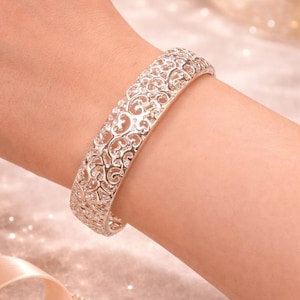 May include: A silver-colored filigree bracelet with an openwork design is displayed on a wrist. The bracelet has an intricate pattern of swirls and curves. The background is a soft, blurred mix of gold and pink tones, with sparkling accents.