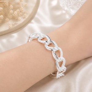 May include: A silver chain link bracelet. The bracelet is made of interlocking oval links. The jewelry is worn on a wrist, and the background is a soft, off-white fabric.