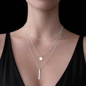 May include: A layered silver necklace set. The top layer features small beads, the middle layer has a small disc pendant, and the bottom layer has a vertical bar pendant. The necklaces are displayed against a black background.