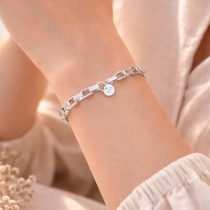 May include: A silver bracelet with a rectangular link chain design. The bracelet features a small, round charm with the text "925" on it. The bracelet is worn on a wrist.