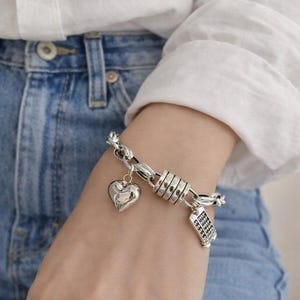 May include: Silver-toned bracelet featuring a heart charm and a charm resembling a calculator. The bracelet has a chain-link design with a series of cylindrical links. The bracelet is worn on a wrist, against a backdrop of a white shirt and blue jeans.