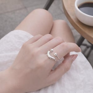 May include: A silver open-style ring featuring a smooth, curved bar on one end and a small, polished spherical bead on the other. The ring has a polished metallic finish and an adjustable band design that remains open at the top for a flexible fit.