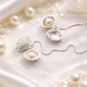May include: A delicate silver necklace featuring three seashell pendants, each holding a lustrous pearl. The necklace rests on a white satin surface, surrounded by scattered pearls, creating an elegant and feminine aesthetic.