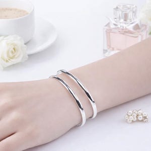 May include: Two polished silver-tone bangle bracelets with a smooth, rounded profile. The bracelets are worn together on the wrist, featuring a reflective metallic finish that catches the light, creating a clean and simple appearance.