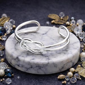 May include: A silver-colored cuff bracelet with a knot design. The bracelet is displayed on a round, marble-like surface, surrounded by decorative crystals and pearls. The bracelet's design features a knot in the center.