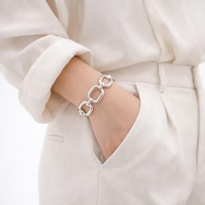 May include: A silver bracelet with a chain link design. The bracelet features a series of rectangular and circular links. The jewelry is worn on a wrist, with the person's hand in a pocket. The background is a white shirt and pants.