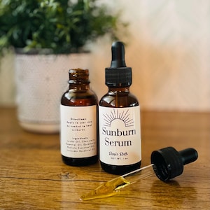 May include: Two brown glass bottles of Sunburn Serum with white labels and black droppers. One bottle has a dropper extended, dispensing the serum. The labels read "Sunburn Serum" and "Ray's Roots". The net weight is 1 fl oz.