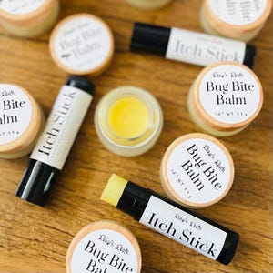 May include: An assortment of natural skincare products, including small jars of Bug Bite Balm and Itch Stick applicators. The products have wooden lids and white labels with black text. A small glass jar contains a yellow balm.