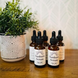 May include: Several brown glass bottles with black droppers are arranged on a wooden surface. Each bottle has a white label with the words "Sunburn Serum" and a sunburst design. A potted plant with green leaves sits to the left.