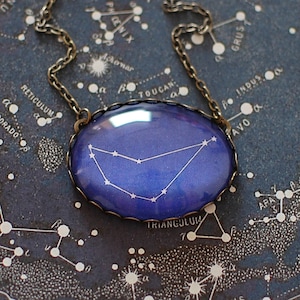 May include: A silver chain necklace with a glass pendant featuring a constellation of stars in a blue and purple gradient against a black background. The pendant is oval shaped and has a silver rim. The necklace is displayed on a vintage star chart.