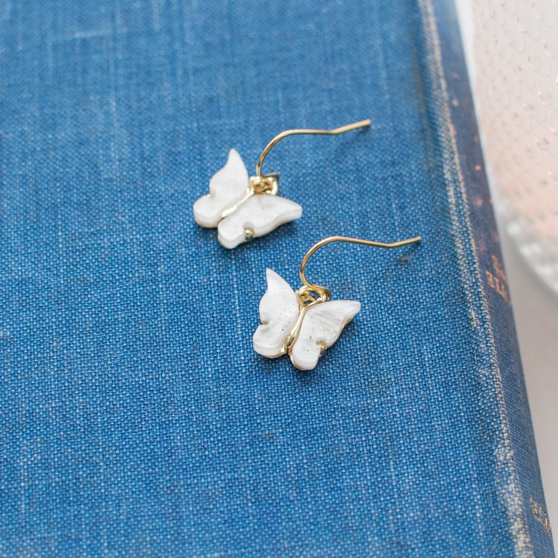 May include: A pair of gold butterfly earrings with white pearl-like wings. The earrings are on a blue fabric background.